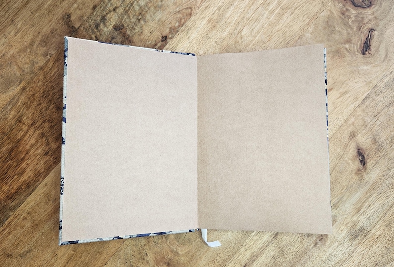 K&ouml;nnte beinhalten: Ein offenes Notizbuch mit leeren, hellbraunen Seiten. Der Einband hat ein Muster in Blau- und Wei&szlig;t&ouml;nen. Ein wei&szlig;es Lesezeichen aus Band ist sichtbar. Das Notizbuch liegt auf einer Holzoberfl&auml;che.
