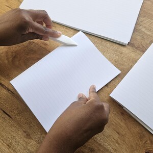 Puede incluir: Una persona usa un plegador para doblar el borde de un papel blanco rayado. Varias pilas de papel blanco est&aacute;n sobre una superficie de madera.