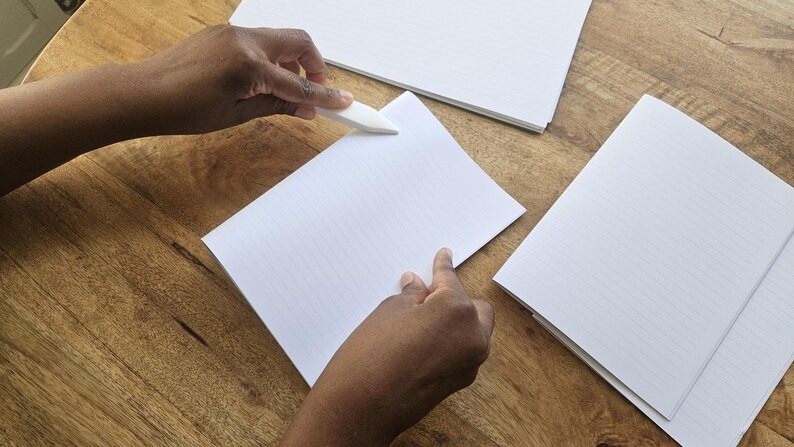 Puede incluir: Una persona usa un plegador para doblar el borde de un papel blanco rayado. Varias pilas de papel blanco est&aacute;n sobre una superficie de madera.