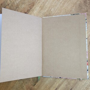 May include: An open journal with blank, tan-colored pages. The journal's cover is visible on the edges, featuring a floral pattern with pink, white, and black elements. A green ribbon bookmark is visible.