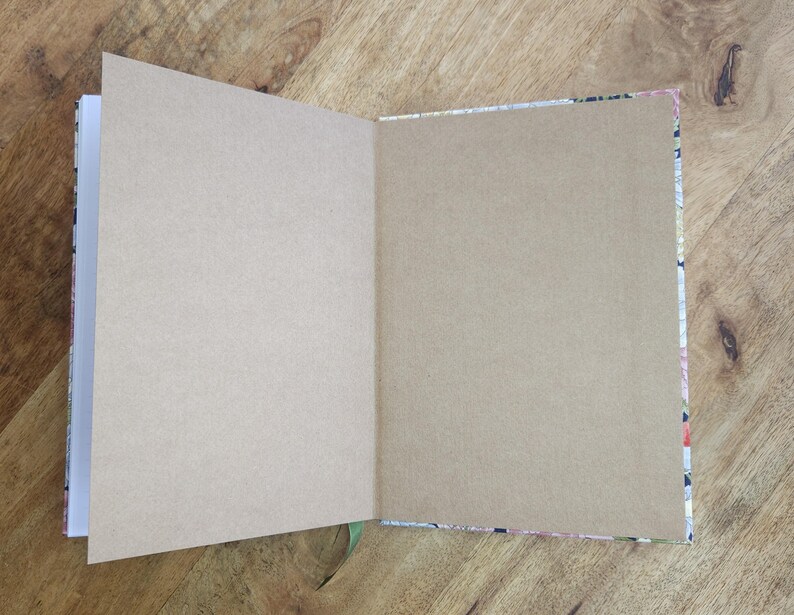 May include: An open journal with blank, tan-colored pages. The journal's cover is visible on the edges, featuring a floral pattern with pink, white, and black elements. A green ribbon bookmark is visible.