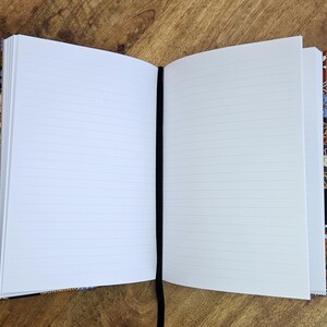Puede incluir: Un cuaderno abierto con p&aacute;ginas blancas rayadas y una cinta negra como marcador. La cubierta tiene un colorido estampado floral. El cuaderno est&aacute; sobre una superficie de madera.