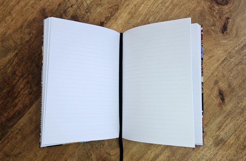 Puede incluir: Un cuaderno abierto con p&aacute;ginas blancas rayadas y una cinta negra como marcador. La cubierta tiene un colorido estampado floral. El cuaderno est&aacute; sobre una superficie de madera.