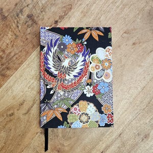 Puede incluir: Un cuaderno negro con un dise&ntilde;o japon&eacute;s colorido. La cubierta presenta un f&eacute;nix, flores y hojas en tonos rojos, naranjas, azules y verdes. Se ve una cinta negra como marcador.