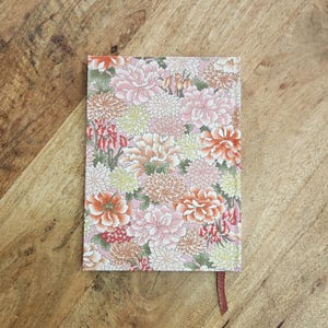 Puede incluir: Un cuaderno con estampado floral con fondo rosa y variedad de flores en tonos rosa, naranja y blanco. El cuaderno tiene un marcador de cinta marr&oacute;n.