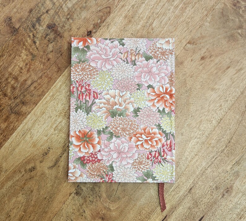 Puede incluir: Un cuaderno con estampado floral con fondo rosa y variedad de flores en tonos rosa, naranja y blanco. El cuaderno tiene un marcador de cinta marr&oacute;n.
