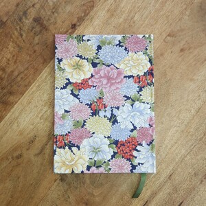 May include: A floral patterned notebook with a dark blue background. The cover features a colorful arrangement of pink, white, yellow, and blue flowers. A green ribbon bookmark is visible. The notebook is on a wooden surface.