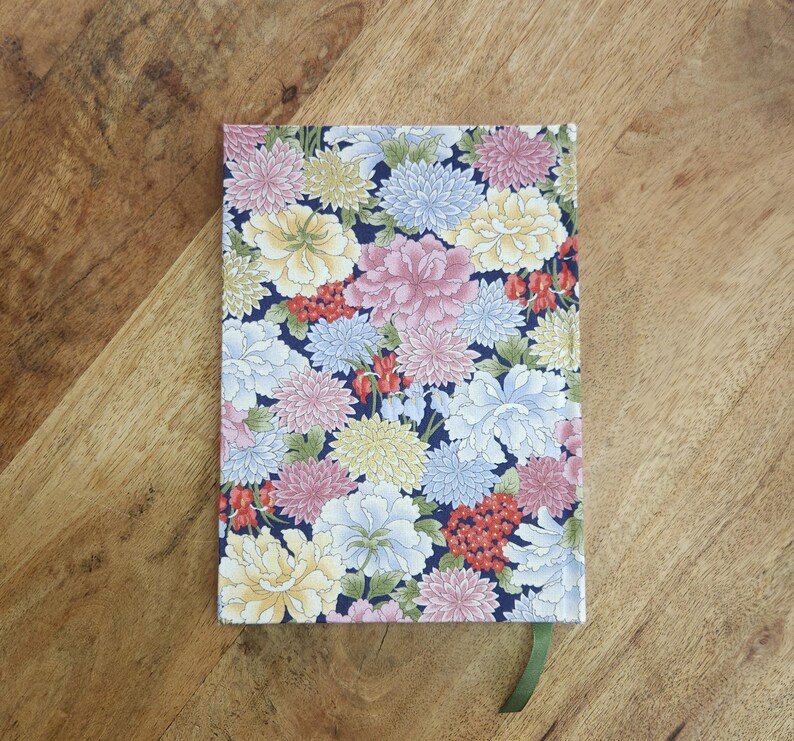 May include: A floral patterned notebook with a dark blue background. The cover features a colorful arrangement of pink, white, yellow, and blue flowers. A green ribbon bookmark is visible. The notebook is on a wooden surface.