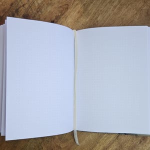 K&ouml;nnte beinhalten: Offenes Notizbuch mit Punktrasterseiten und einem cremefarbenen Schleifen-Lesezeichen. Das Notizbuch liegt offen auf einer Holzoberfl&auml;che. Der Einband hat ein schwarz-wei&szlig;es Muster.