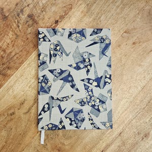 K&ouml;nnte beinhalten: Ein rechteckiges Notizbuch mit hellgrauem Einband, das ein Muster aus Origami-Kranichen und geometrischen Formen in Blau- und Wei&szlig;t&ouml;nen zeigt. Ein wei&szlig;es Lesezeichenband ist sichtbar.