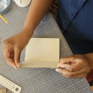 Puede incluir: Una persona trabaja en un proyecto de manualidades, posiblemente encuadernaci&oacute;n. Est&aacute; doblando un trozo de cart&oacute;n marr&oacute;n claro. Otros materiales incluyen una regla, pegamento y una esterilla de corte.