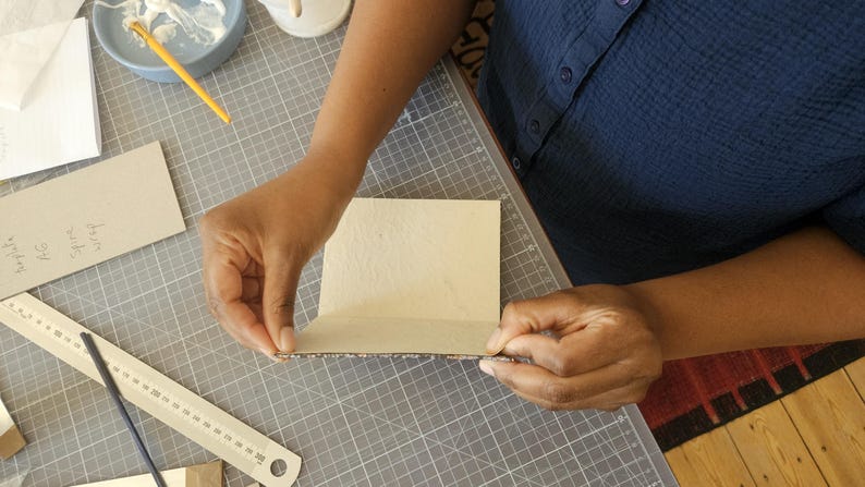 Puede incluir: Una persona trabaja en un proyecto de manualidades, posiblemente encuadernaci&oacute;n. Est&aacute; doblando un trozo de cart&oacute;n marr&oacute;n claro. Otros materiales incluyen una regla, pegamento y una esterilla de corte.