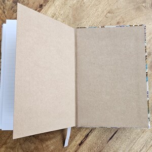 May include: An open notebook with lined pages and a kraft paper cover. The right-hand page is kraft paper, and the left-hand pages are lined. The notebook is on a wooden surface, and a white ribbon bookmark is visible.
