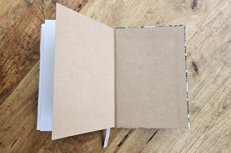 May include: An open notebook with lined pages and a kraft paper cover. The right-hand page is kraft paper, and the left-hand pages are lined. The notebook is on a wooden surface, and a white ribbon bookmark is visible.