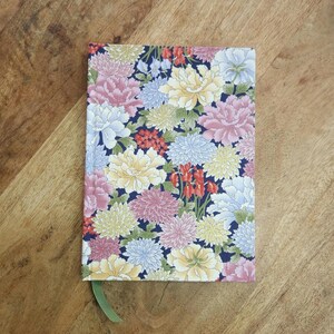 May include: A floral patterned journal with a dark blue background. The cover is covered in pink, yellow, white, and red flowers. A green ribbon bookmark is visible. The journal is rectangular and sits on a wooden surface.