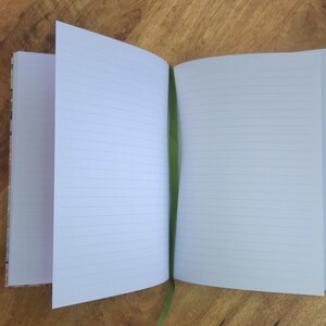 May include: An open notebook with lined pages and a green ribbon bookmark. The notebook's cover has a colorful floral pattern. The pages are white with horizontal lines, perfect for writing or sketching. The notebook is on a wooden surface.