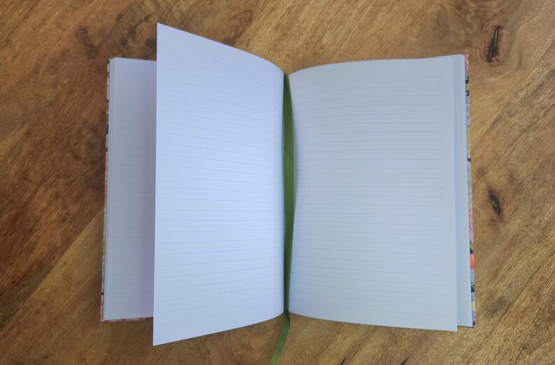 May include: An open notebook with lined pages and a green ribbon bookmark. The notebook's cover has a colorful floral pattern. The pages are white with horizontal lines, perfect for writing or sketching. The notebook is on a wooden surface.