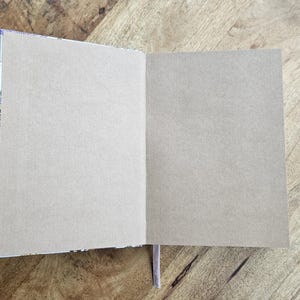 May include: An open notebook with blank, light-colored pages. The notebook's spine has a decorative pattern, and a thin, light purple ribbon bookmark is visible. The notebook is on a wooden surface.