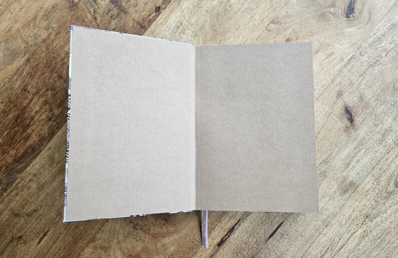 May include: An open notebook with blank, light-colored pages. The notebook's spine has a decorative pattern, and a thin, light purple ribbon bookmark is visible. The notebook is on a wooden surface.