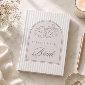 Puede incluir: Un libro de tapa dura con un patr&oacute;n de rayas y el texto "Letters to the Bride". La portada presenta dos dise&ntilde;os en forma de coraz&oacute;n con las letras "N" y "H". Tambi&eacute;n se ve un bol&iacute;grafo dorado, anillos en un plato y una vela encendida.