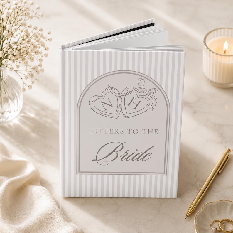 Puede incluir: Un cuaderno de tapa dura con un patr&oacute;n de rayas y el texto "Letters to the Bride". La portada presenta un dise&ntilde;o con dos medallones en forma de coraz&oacute;n con las letras "N" y "H". Un bol&iacute;grafo dorado y anillos est&aacute;n al lado.