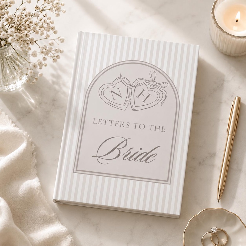 Puede incluir: Un libro de tapa dura con un patr&oacute;n de rayas y el texto "Letters to the Bride". La portada presenta dos dise&ntilde;os en forma de coraz&oacute;n con las letras "N" y "H". Tambi&eacute;n se ve un bol&iacute;grafo dorado, anillos en un plato y una vela encendida.