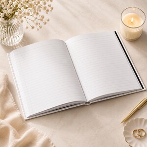Puede incluir: Un cuaderno abierto con p&aacute;ginas rayadas y una cubierta blanca. Una vela encendida en un portavelas de vidrio, un bol&iacute;grafo dorado y un plato peque&ntilde;o con dos anillos de oro tambi&eacute;n est&aacute;n presentes. Un peque&ntilde;o jarr&oacute;n de vidrio con flores blancas est&aacute; en el fondo.