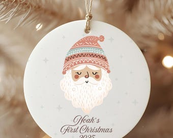 May include: A round Christmas ornament featuring a Santa Claus design. The Santa has a pink and blue hat, a white beard, and closed eyes. The ornament reads "Noah's First Christmas 2025" in a script font. A beige string is attached for hanging.