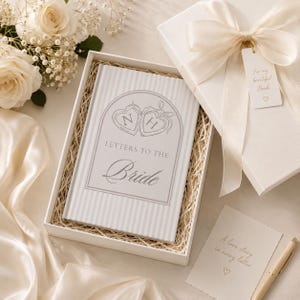 Puede incluir: Una caja de regalo blanca con un lazo contiene un libro titulado "Cartas a la novia". El libro tiene una cubierta a rayas con un dise&ntilde;o de coraz&oacute;n. Una etiqueta dice "Para mi hermosa novia". Tambi&eacute;n hay un bol&iacute;grafo y una tarjeta.