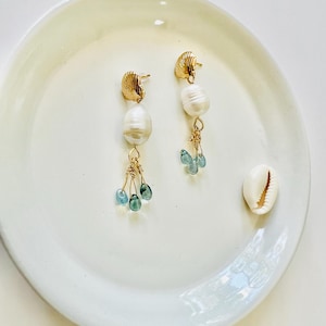 May include: Gold-toned earrings with a seashell design at the top, followed by a large white pearl and a cluster of small, blue-green beads. The earrings are displayed on a white plate, with a small seashell to the side.