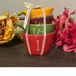 May include: A stack of Rae Dunn ceramic measuring cups in red, green, burgundy, and yellow. Each cup is labeled with a measurement: 1, 1/2, 1/4. The cups are tied together with a white ribbon that reads "Rae Dunn".