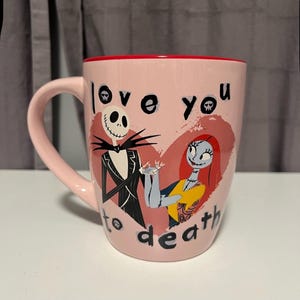 May include: Pink ceramic mug featuring Jack Skellington and Sally from The Nightmare Before Christmas, with the words "love you to death." The mug has a red interior and a curved handle.