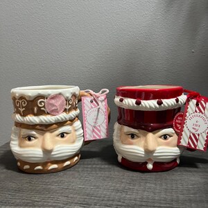 May include: Two ceramic mugs shaped like Santa Claus faces. One mug is brown and white, the other is red and white. Each mug has a handle and a tag that says "Peppermint Fine".