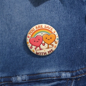 May include: A round button with the text "YOU ARE SAFE WITH ME" encircling a rainbow and two cartoon hearts holding hands. The hearts are pink and orange with smiling faces. The button is on a blue denim jacket.