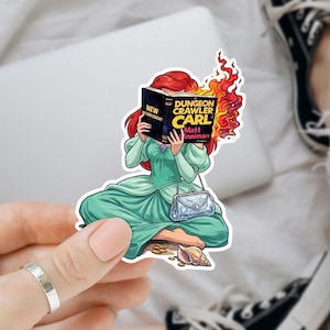 A person is holding a sticker of a cartoon girl reading a book, with a pair of black and white sneakers visible in the background.