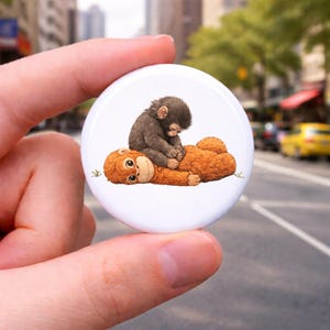 May include: A white button featuring an illustration of two orangutans. One is lying down, and the other is sitting on top, appearing to groom the other. The orangutans are brown and orange, with a detailed, cartoon-like style.