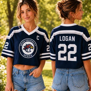 A young woman wearing a navy blue crop top with a white and navy blue design, a white "C" on the front, and the name "Logan" and the number "22" on the back. She is also wearing denim shorts.