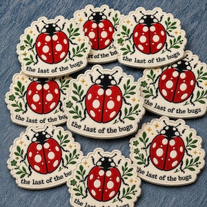May include: A collection of embroidered patches featuring a red ladybug with white spots and a black head. Each patch is surrounded by a cream-colored border with green leaves and the text "the last of the bugs".