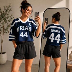 May include: A navy blue cropped jersey with white stripes on the sleeves, featuring the text BRIAR and the number 44 on the front, and the text GRAHAM and the number 44 on the back. The jersey is paired with solid black athletic shorts.