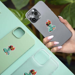 A hand holding a smartphone with a sticker on the back cover, and there are other cases with stickers on them, placed on a light green surface.