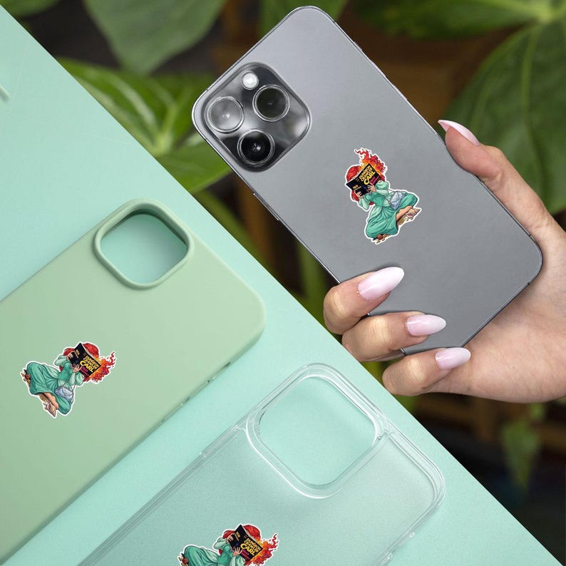 A hand holding a smartphone with a sticker on the back cover, and there are other cases with stickers on them, placed on a light green surface.