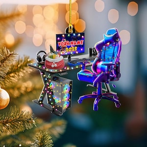 May include: A miniature gaming setup ornament featuring a computer monitor with the text "YOUR NAME 2025", a gaming chair, and a desktop computer. The desk and computer are decorated with colorful lights. A red bag with gifts sits on the desk.