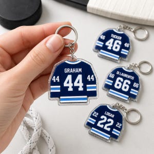 May include: A collection of hockey jersey-shaped keychains. The keychains are navy blue with white accents, featuring player names and numbers. One keychain displays "GRAHAM 44", while others show "TUCKER 46", "DI LAURENTIS 66", and "LOGAN 22".