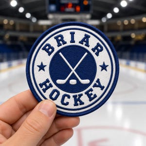 May include: A round, embroidered patch with a blue and white design. The patch features the words "BRIAR HOCKEY" in white lettering around a central blue circle with crossed hockey sticks and a puck. Two stars are on either side.