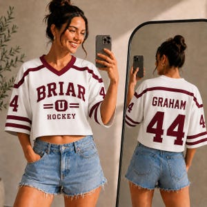 May include: A white cropped hockey jersey with maroon accents, featuring a V-neck, short sleeves, and the text BRIAR HOCKEY with a U logo on the front. The back displays the name GRAHAM and the number 44 in maroon, with the number 4 on the sleeves.