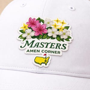 May include: White baseball cap featuring a floral patch with pink, white, and yellow flowers, green leaves, and the text "MASTERS AMEN CORNER." A yellow map with a red flag is also on the patch.