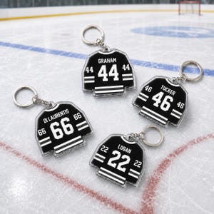 May include: Four hockey jersey-shaped keychains. Each keychain is black with white stripes and numbers. The names and numbers on the jerseys are Graham 44, Tucker 46, Di Laurentis 66, and Logan 22. The keychains are on a hockey rink surface.