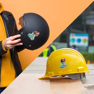 a person holding a black motorcycle helmet and a yellow construction helmet with stickers on them, both placed on a wooden surface.