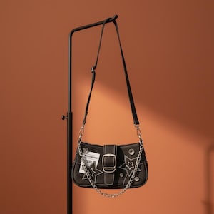 Puede incluir: Bolso de hombro de mezclilla negra con parches de estrella y detalles de cadena plateada. El bolso tiene un cierre de hebilla y una pequeña etiqueta. El bolso está suspendido de un soporte de metal negro sobre un fondo naranja.