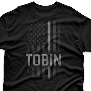 May include: Black t-shirt featuring a distressed American flag design with the name "TOBIN" in a bold, gray font. The flag design is vertically oriented, with the stars and stripes visible. The shirt is made of a soft material.